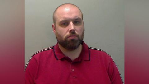 A bald man with a brown beard looks into the camera. He is wearing a red polo shirt.