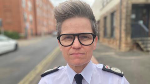 Head and shoulders shot of Rachel Swann - she is wearing her police uniform, has thick black-rimmed glasses and has a large fringe-type hairstyle. 