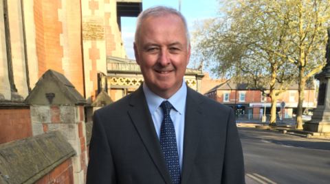 Staffordshire police, fire and crime commissioner Ben Adams. He is wearing a grey suit and pale blue shirt with navy blue tie, and is stood outside Burton Town Hall.