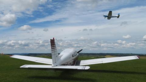 A single engine white and red plane sits on the grass as another blue and white aircraft flies above it in cloudy skies with blue patches