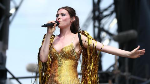 Sophie Ellis-Bextor wearing a gold outfit on stage, covered in gold streamers. she is singing into a microphone and has an outstretched arm.
