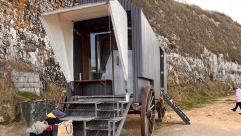 An exterior image of a free-to-use sauna in Margate which has attracted visitors from all over the south east. The sauna is elevated above the ground and has four steps up into it. Above the entrance there is a white tarpaulin which extends out. Either side of the sauna are two wheels; two larger wheels at the front and two smaller wheels at the back.