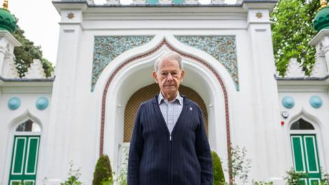 An elderly man wearing a grey shirt and dark blue cardigan. He is facing the camera and stood outside a white mosque