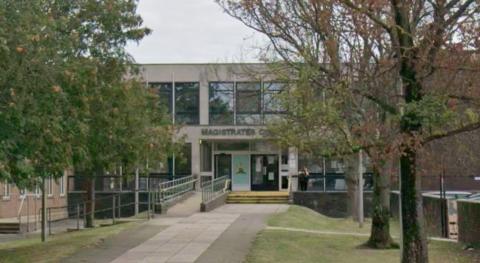 A single-storey building set back from the street, with a sign above the entrance reading “Magistrates Court”. A paved walkway with trees on either side leads directly to the front doors, flanked by metal railings on a sloped access ramp.