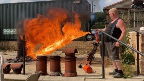 Claughton uses the device, creating a large burst of bright orange fire. He is standing on a paved area next to a brick wall and metal handrail, wearing a sleeveless top, shorts, and sandals. To his left are several cylindrical metal objects arranged in a row on the ground. Behind the flaming area, there is a large green shipping container and some scattered equipment or debris on the ground. A plume of dark smoke rises into the sky from behind the container.