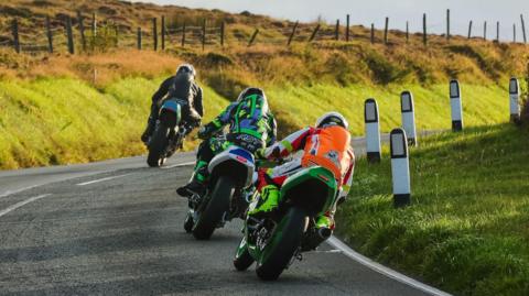 The backs of three racers on bikes rounding a bend on the Mountain Road. There is a grass verge and a farm fence on the outside of the bend on the left and reflective bollards on a grassed area on the right.