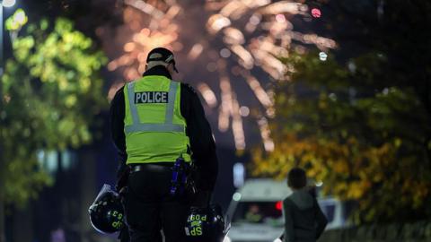 A police officer wearing a high-visibility vest stands on a street at night holding two helmets, with fireworks lighting up the sky in the background