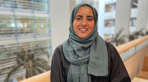 A woman wearing a black top and a green/blue headscarf. She is stood on a balcony with palm trees and office space in the background.
