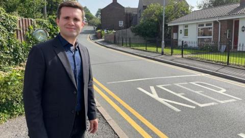 A man with short brown hair, wearing a dark jacket and blue shirt standing next to a road. 