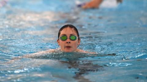 A woman with dark hair in goggles is swimming in an indoor pool