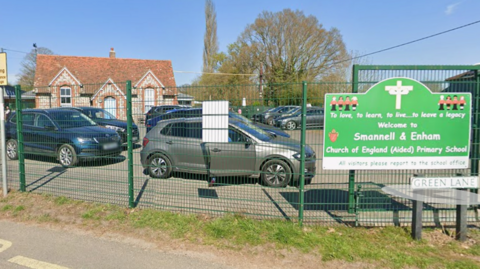 A small bred brick building, with around six cars parked in a car park in front of it, and a green metal fence and gate with a green sign on it saying 'Welcome to Smannell and Enham Church of England primary school"