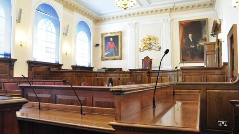 A picture of the inside of Guernsey's States Chamber with desks and microphones in the foreground and the States crest on the wall in the background with two large painted portraits on either side.