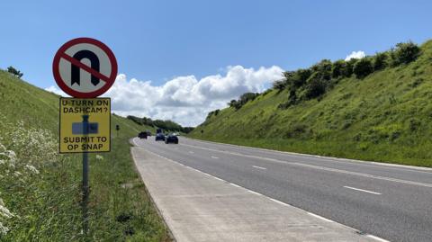 A30 Hayle bypass in Cornwall. Vehicles are travelling along the road. Green banks lined either side of the carriageway. U-turn signage has been installed, including an Operation Snap sign which says "U-turn on dashcam? Submit to Op Snap"
