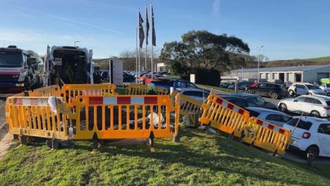 A site on an industrial estate. In the middle is a a yellow temporary barrier on a grass verge. Cars and lorries can be seen to side the side and in the background.