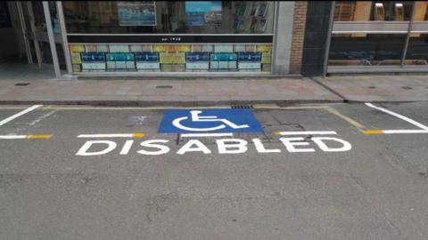 A parking space on a road with a logo of a person in a wheelchair and the word DISABLED. Above the pavement are two glass-fronted shops.