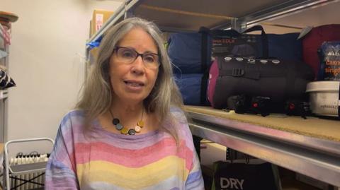 A woman with glasses and a striped top and a chunky necklace. Behind her are shelves with camping accessories.
