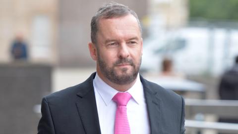 Andrew Polson, a man with thin grey/brown hair and a beard.  He is walking down the street while wearing a dark suit and pink tie.