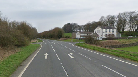 A quiet road with three lanes, one going forward, one to turn right and another to go in the opposite direction. In the top right is a pub or a hotel with a white facade.