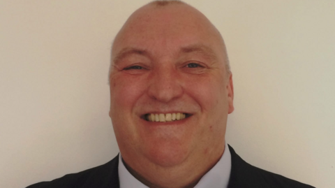 Headshot of Mark. He is smiling at the camera, in front of a pale background. He has short shaved head. 