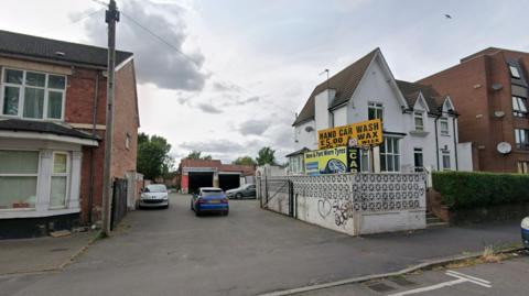 A flat tarmacked area with at least three cars on it is photographed. A yellow sign next to the area reads: "Hand car wash and wax £5."