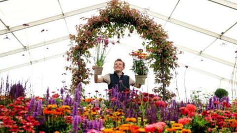 A man holding up two pots of flowers under an arch of flowers inside a marquee. There are many colourful flowers in the foreground.