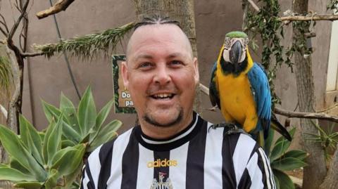 Anthony Robson is wearing a black and white Newcastle top with a parrot on his shoulder. He is smiling to the camera and has a chin beard. 