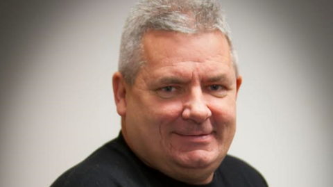 Ollie Green has grey hair, is smiling at the camera  and is wearing a black t-shirt.