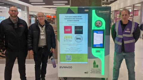 Three people stand inside a busy shopping centre beside a bright EcoVend reverse‑vending machine, which collects empty plastic bottles and cans. The group appear to be staff or project partners, wearing uniforms and lanyards as they pose for the photo. Behind them, high‑street shops and food outlets form the backdrop, emphasising the machine’s prominent position in a public retail space. The EcoVend unit displays messaging about turning recyclables into in‑store rewards, along with logos of participating local businesses.