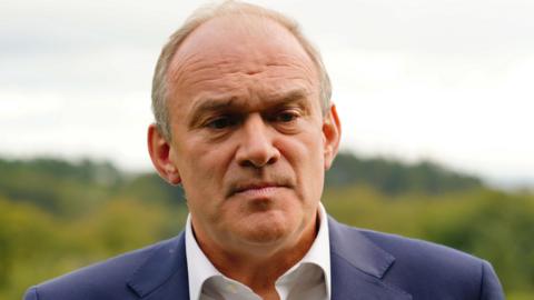 Lib Dem leader Sir Ed Davey wearing a white shirt, blue suit and neutral expression