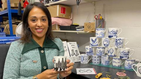 A woman with shoulder length dark hair sits beside a tower of 14 decorated china cups. The woman is holding one of the cups which has a black and white design. She is wearing a green and white speckled dress with large buttons and dark green trim. In the background, boxes, scissors and stationary can be seen.