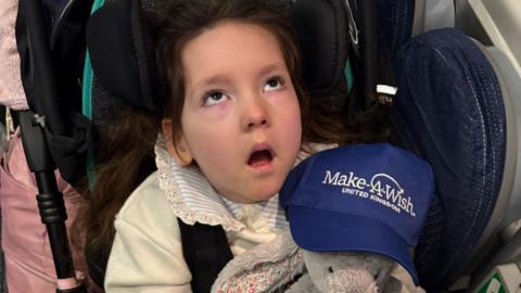 An image of a young girl looking upwards with a cuddly toy on her lap. A hat wiith the words 'Make-A-Wish' United Kingdom'  is also seen in the photograph 