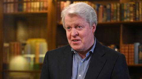 Earl Spencer is photographed in a dark jacket and blue and white checked shirt that is open at the neck. He has greying hair. In the background are blurred shots of bookcases filled with books.