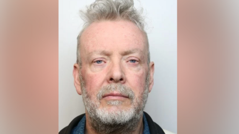 A police mugshot of a man - Sean Morris from Massinger Walk in Swindon - with grey/blue eyes, grey hair, moustache and beard and wearing a black jacket looking directly into the camera. 