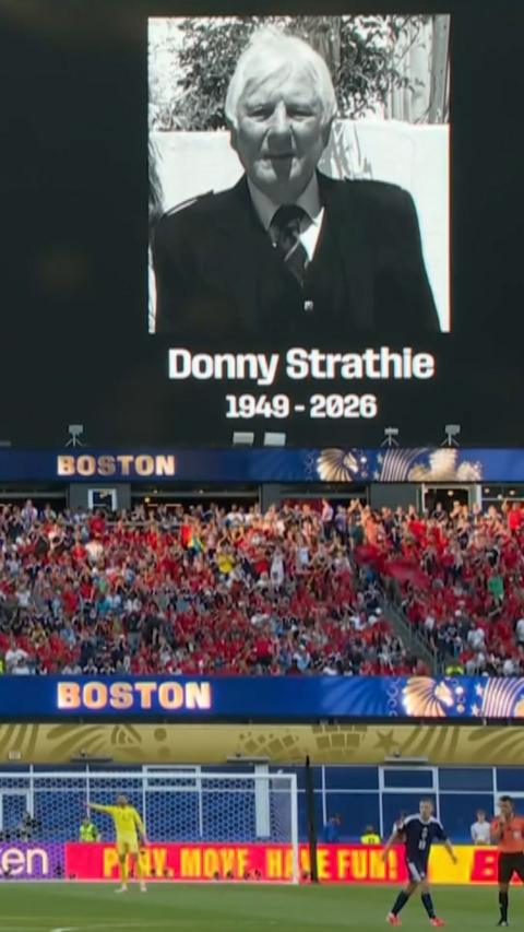 A black and white photo of Donny Strathie with 1949-2026 is on the screen above a packed football pitch.