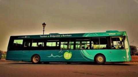 A green single-decker Somerset bus against a grey sky. It has "Taunton, Bridgwater and Burnham" written on it