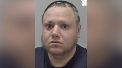 A man with short dark hair and a dark blue T'shirt in a police mugshot.