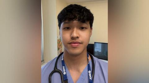 A young male doctor wearing a purple uniform with a stethoscope around his neck. He is looking into the camera