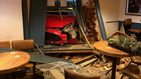 A red car has crashed through the window and wall of a pub - there is significant damage to the walls, and the pub is strewn with debris. Tables and chairs can be seen in the establishment, some with Christmas flower arrangements on top. The car is badly damaged.