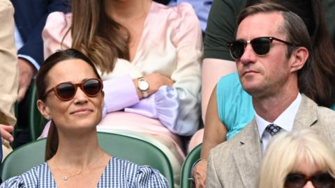 A picture of James and Pippa Matthews, both wearing sunglasses, sat at Wimbledon. James is wearing a stone-coloured suit and Pippa is wearing a blue and white dress.