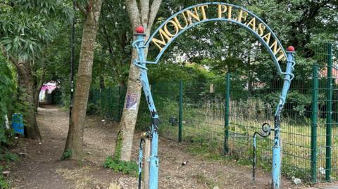 A dilapidated looking sign in front of a wooded path next to a fence