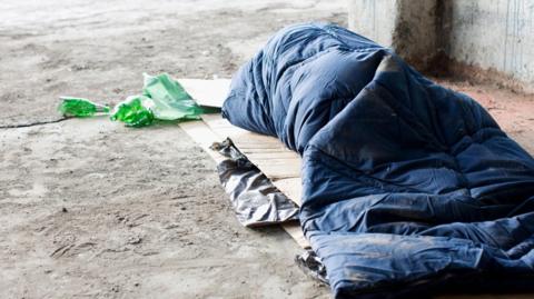 A homeless person sleeping in sleeping bag on cardboard. They are in a blue sleeping bag on top of some cardboard and there is a green food packet on the floor.