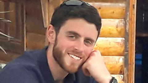 PC Andrew Harper smiles into the camera with his chin resting on his closed left hand. He is wearing a navy top and has a pair of sunglasses perched on the top of his head. He has dark brown short hair and a brown short beard