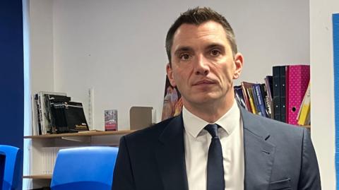 Dominic Wilkes is looking at the camera as he stands in a classroom. He is wearing a navy jacket and tie and a white shirt. He has short black hair, brown eyes. There are chairs on top of tables behind him.