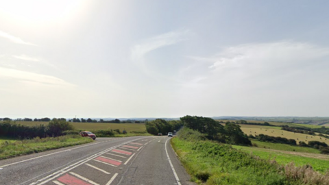 Google Street View of the A37 - a wide, two-way, tarmac road passing through picturesque countryside with far-reaching views of farm fields, meadows and hedgerows.
