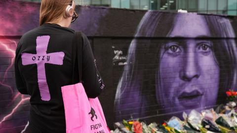 A young woman with a large purple cross and the word Ozzy on the back of her black top, facing a purple mural of Ozzy's face, with a line of flowers below