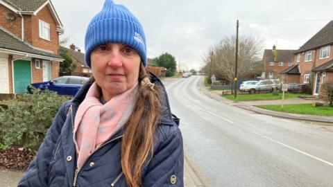 A woman with a blue hat, pink scarf and a blue coat standing by the side of a long main road