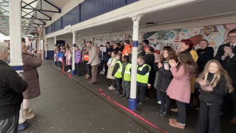 A group of children and adults are gathered on a train station platform unveiling the mural. Some people are taking photos while others clap, with a colourful mural providing a bright backdrop behind the crowd.