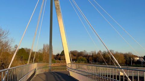 General view of a bridge in Worcester which connects Gheluvelt Park to St John's. The bridge curves to the right with wires coming award from a central silver spire. 