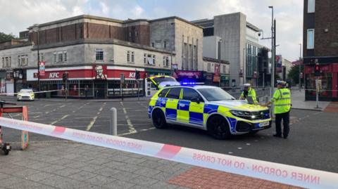 Police cordon with two police cars inside the cordon at a large city centre junction.  Two police officers stand at police car seen to the right.
