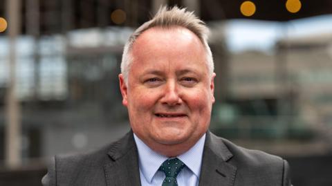 Darren Millar stood in the centre of the photograph wearing a grey suit, green tie and blue shirt.
